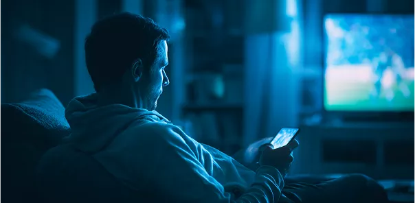 Người đàn ông ngồi trên ghế sofa, xem TV và sử dụng điện thoại di động trong phòng tối.